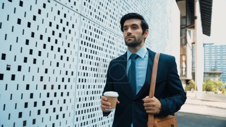 Photo for Skilled business man walking in suit outfit while holding coffee cup. Professional manager standing near architectural building. Going to working in morning, seeking for successful job. Exultant. - Royalty Free Image