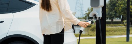 Photo for Young woman recharge EV car battery at charging station connected to power grid tower electrical industrial facility as electrical industry for eco friendly vehicle utilization. Panorama Expedient - Royalty Free Image