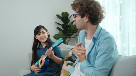 Back view of happy father looking at smart daughter playing ukulele warmly and singing song. Happy dad motivated and encouraged american school girl enjoy playing guitar. Family recreation. Pedagogy.