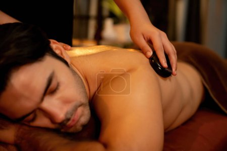 Photo for Hot stone massage at spa salon in luxury resort with warm candle light, blissful man customer enjoying spa basalt stone massage glide over body with soothing warmth. Quiescent - Royalty Free Image