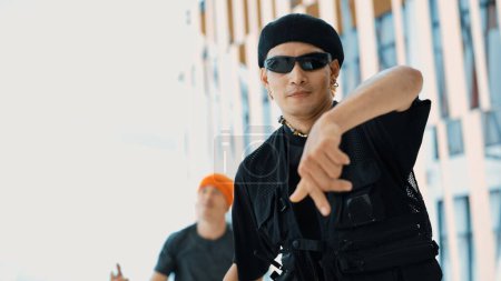 Asian hipster looking at camera while pose at camera while friend dancing behind at sky scrapper. Group of street dancer perform break dancing while looking at camera. Outdoor sport 2024. Endeavor.