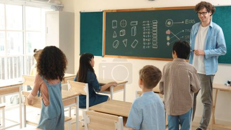 Diverse elementary student walking in classroom while teacher looking at child. Cute children attend classroom while professional instructor prepare for teaching and standing at blackboard. Pedagogy.