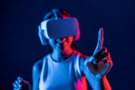 Foto de Soporte femenino inteligente con auriculares VR rodeados de luz de neón cyberpunk que conectan tecnología de comunidad ciberespacial metaversa y futurista. Mujer usando el dedo señalando objeto virtual. Alucinaciones. - Imagen libre de derechos