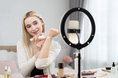 Young woman making beauty and cosmetic tutorial video content for social media. Beauty blogger smiles to camera while showing how to apply beauty skincare to audience or followers. Blithe