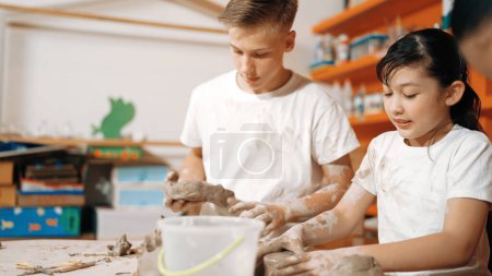 Photo for Caucasian boy modeling bowl of clay while cute asian girl working on dough at pottery workshop. Skilled highschool student playing on dough while wear muddy shirt at art lesson. Creative. Edification. - Royalty Free Image