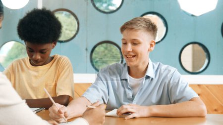 Photo for Young smart teenager writing and talking about topic at classroom. Diverse highschool student taking a note in other notebook while sitting with multicultural friends. Creative education. Edification. - Royalty Free Image