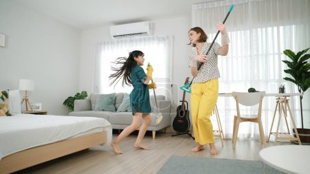 Photo for Caucasian mom and asian child doing housework together and dancing to music. Attractive mother and daughter cleaning modern bedroom while sing along and jump to relaxing music. Lifestyle. Pedagogy. - Royalty Free Image