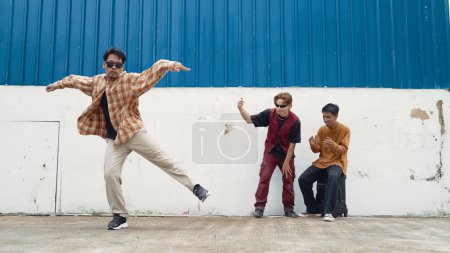 Foto de Joven bailarina callejera bailando en estilo hip hop con amigos multiculturales animando detrás de la pared. Atractivo hombre guapo estirar los brazos en la calle con fondo azul. Deporte al aire libre 2024. Endeavor. - Imagen libre de derechos