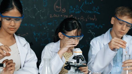 Schlaue Teenager werfen Proben ins Mikroskop, während Mädchen an der Tafel mit Chemie-Theorie sorgfältig inspizieren. Fachwissenschaftler gießen chemisches Produkt in Röhre und bereiten sich auf Experimente vor. Erbauung.