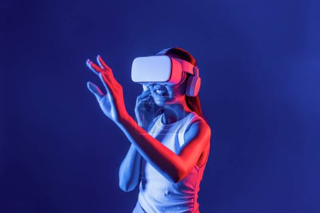 Foto de Mujer inteligente de pie con auriculares VR rodeados de luz de neón cyberpunk que conectan tecnología de comunidad del ciberespacio metaversa y futurista. Mujer usando mano tocando un objeto virtual. Alucinaciones. - Imagen libre de derechos