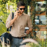 Young handsome man walk out of coffee shop with cafe garden, holding a coffee cup and a shopping bag while talking on the phone. Modern happy carefree with sunglasses lifestyle. Expedient