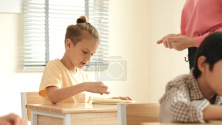 Photo for Beautiful teacher explaining test while caucasian boy doing classwork and listening explanation. Cute child looking at teacher while taking a notes or writing answer in paper at classroom. Pedagogy. - Royalty Free Image
