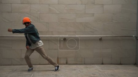 Photo for Asian smart hipster show energetic break dance with gray background at building while wearing headphone and stylish cloth and listen hip hop song. Outdoor sport 2024. Hip hop movement. Sprightly. - Royalty Free Image