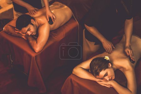 Téléchargez les photos : Massage aux pierres chaudes au salon de spa dans un complexe de luxe avec lumière chaude aux bougies, un couple heureux profitant d'un massage aux pierres de basalte spa glisse sur le corps avec une chaleur apaisante. Tranquillité - en image libre de droit