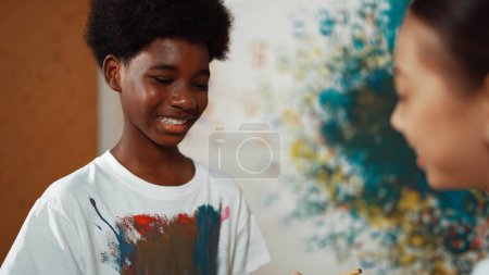 Téléchargez les photos : Souriant divers enfants en utilisant la couleur peinte au pinceau sur chaque chemise blanche à un mur coloré dans la leçon d'art. Représenter l'échange d'expériences, apprendre les uns les autres. Édification. - en image libre de droit