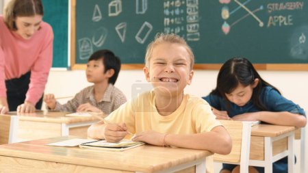Téléchargez les photos : Enfant intelligent caucasien souriant à la caméra tout en faisant du travail en classe tout en étudiant multiculturel heureux faire des tests ou note d'écriture en classe au primaire. Concept d'éducation. Pédagogie. - en image libre de droit
