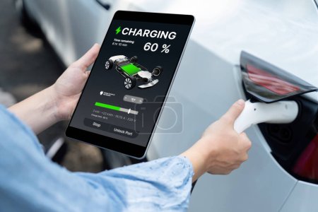 Hand insert EV charger plug into electric vehicle to recharge EV car, battery status display on tablet EV application. Future alternative clean and sustainable energy for transportation. Perpetual