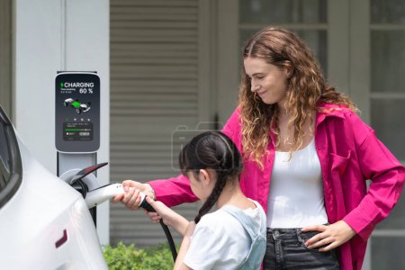 Photo for Happy little young girl learn about eco-friendly and energy sustainability as she help her mother recharge electric vehicle from home EV charging station. EV car and modern family concept. Synchronos - Royalty Free Image