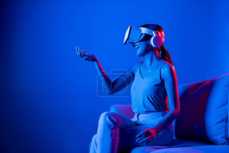 Foto de Las hembras inteligentes sentadas en un sofá rodeado de auriculares VR de neón conectan tecnología de comunidad del ciberespacio metaversa y futurista, observando e interactuando con el objeto virtual con la mano. Alucinaciones. - Imagen libre de derechos