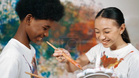 Photo for Smiling diverse children using paint brush painted color on each other white shirt shirt at colorful stained wall in art lesson. Represent exchanging experience, learning each other. Edification. - Royalty Free Image