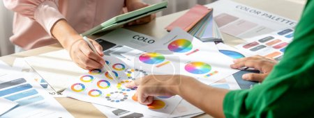 Fachkundiger Architekt sucht Daten vom Tablet, während er eine passende Farbe aus dem Farbrad am Tisch mit Farbpalette und Dokumentenstreuung auswählt. Kreatives Gestaltungskonzept. Bunt gemischt.