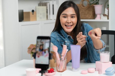 Joven hermosa asiática mostrando maquillaje cosmético lápiz labial mate pastel prueba de color en la colección bestseller en brazo prueba promoción en las redes sociales grabación en línea smartphone en la habitación moderna. Estratagema.