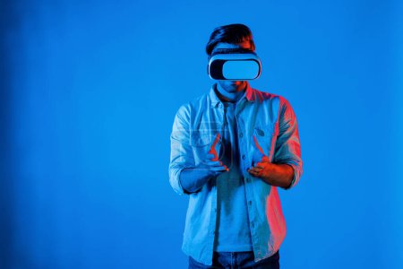 Téléchargez les photos : Homme intelligent caucasien tenant et geste émouvant tout en utilisant le masque VR. Une personne heureuse utilisant un casque et un masque tout en entrant dans le monde virtuel ou métaverser avec un fond lumineux au néon. La technologie. Déviation. - en image libre de droit