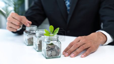 Businessman put coin to money saving glass jar at his office as sustainable money growth investment or eco-subsidize. Green corporate promot and invest in environmental awareness. Gyre