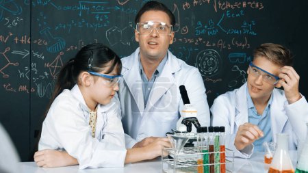 Kreativer Lehrer zeigt Chemie auf Tafel, während er mit einem Gymnasiasten am Tisch mit Mikroskop und Reagenzglas mit farbiger Flüssigkeit spricht. Junge Studentin im Laborkittel im MINT-Unterricht. Erbauung.