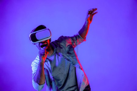 Excited man wearing VR goggle to exploring and enter in virtual program. Skilled gamer touching in metaverse while wearing casual cloth and standing at neon light background. Lifestyle. Deviation.