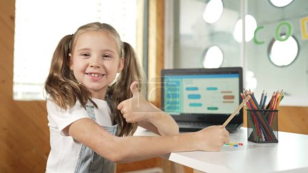 Chica sonriente usando el ordenador portátil mientras mira y mostrar el pulgar hacia arriba a la cámara. Lindo niño que usa auriculares mientras trabaja mediante el uso de código de escritura portátil en la educación de tecnología STEM. Aprendizaje online. Erudición.