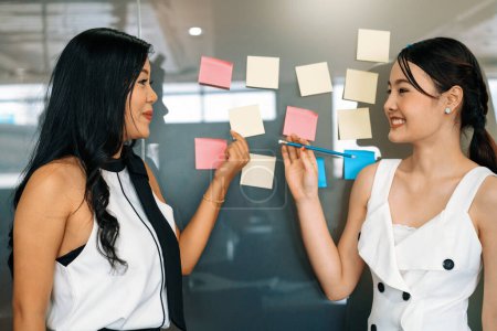 Photo for Two young beautiful Asian businesswomen talk and discuss creative post on the window. It is project reminder and thinking idea notes. uds - Royalty Free Image