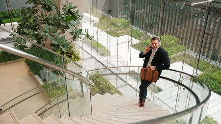 Draufsicht auf einen kaukasischen Geschäftsmann, der mit Kopfhörer entspannende Musik hört, während er an Wendeltreppen eines modernen Gebäudes steht. Projektmanager hören gerne Lieder, während sie zu Reimen übergehen. Urbane Pflanzen.
