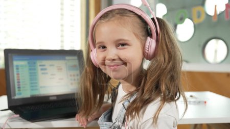 Smiling smart caucasian girl show system programing or coding program in STEM technology education. Child wearing headphone while working by using laptop writing code and look at camera. Erudition.
