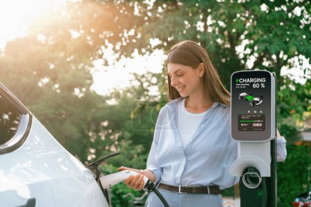 Photo for Young woman recharging battery for electric car during road trip travel EV car in natural forest or national park with sunrise lighting. Eco friendly travel during vacation and holiday. Exalt - Royalty Free Image