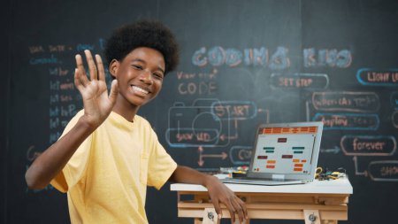 Téléchargez les photos : Le garçon africain intelligent utilise un ordinateur portable pour programmer et se retourner pour saluer la main. Mignon lycéen stident codage ingénierie invite ou système de programme au tableau noir dans la classe de technologie STEM. Édification - en image libre de droit