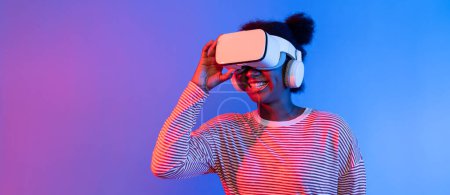 Téléchargez les photos : Une jeune Africaine enthousiaste et souriante portant un casque VR surprenant à travers le monde métaverse dans un studio lumineux rose fluo et bleu vibrant qui apprend la technologie du fond 3D cyberespace. Contrivance. - en image libre de droit