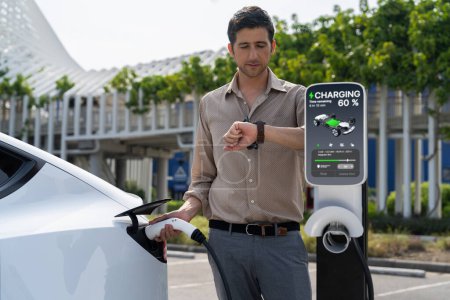 Photo for Young man checking time on smartwatch while EV charger to recharging battery from charging station in city mall parking lot. Rechargeable EV car for sustainable eco friendly urban travel. Expedient - Royalty Free Image