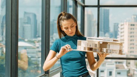 Photo for Young smart architect engineer holds architectural model while inspect house model. Professional interior designer checking house construction while standing near window with city view. Tracery - Royalty Free Image