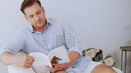 Mann mittleren Alters sitzt auf dem Sofa lesen Fabel Buch erhöhen das Wissen über soziale und Phantasie für Beruf oder erziehen Kinder zu einem guten Erwachsenen als kluger Vater. Konzept des lebenslangen Lernens. Divergenz.