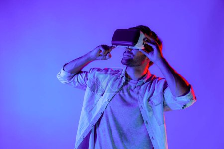Photo for Caucasian man enter metaverse by using virtual digital goggles while standing look in cyberspace display virtual world. Sport gamer with casual cloth move gesture at neon background. Tech. Deviation. - Royalty Free Image