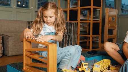 Photo for Caucasian girl sits at sand box while placed car model at slope toy. Diverse children playing plastic model while sitting at school in break time at play room. Creative activity concept. Erudition. - Royalty Free Image