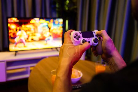 Fermez un joueur mains homme de jeu vidéo à la télévision en utilisant le contrôle joystick avec écran de jeu de combat flou avec collation et boissons à la décoration avec néon blub lumière dans la salle de séjour confortable. Postuler.