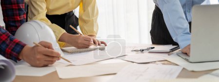 Photo for Professional engineer draws a blueprint on meeting table with blueprint and equipment scattered around while project manager using laptop analysis data at meeting room. Closeup. Delineation. - Royalty Free Image