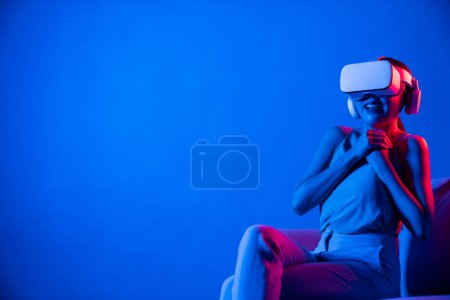 Foto de Las hembras inteligentes sentadas en un sofá rodeado de auriculares VR de neón conectan tecnología de comunidad ciberespacial metaversa y futurista. Mujer elegante emocionado y emocionalmente ver películas. Alucinaciones. - Imagen libre de derechos