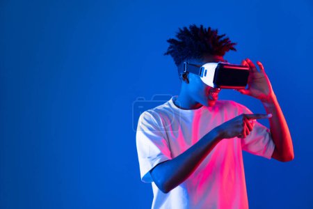 Téléchargez les photos : Étonné jeune afro-américain regardant à travers VR tenant hologramme objet intéressant sur le mur néon rose bleu au monde métaverse reliant la technologie numérique futuriste réalité virtuelle. Contrivance. - en image libre de droit