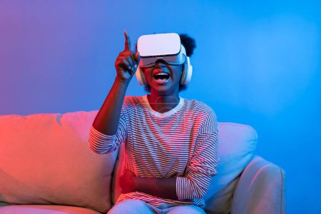 Photo for Surprised African American woman watching film through VR in fantastic 3D cartoon movie technology virtual reality metaverse neon lighting journey new world at living room sitting sofa. Contrivance. - Royalty Free Image