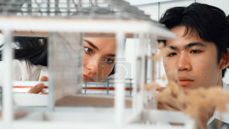 Close up of cooperative professional engineer working together to measuring house model structure. Young beautiful architect measure house model while male engineer support performance. Immaculate.