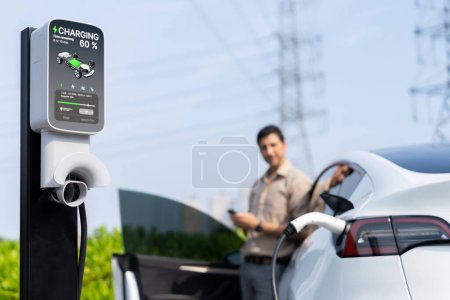 Photo for Man pay for electricity with smartphone while recharge EV car battery at charging station connected to power grid tower electrical as electrical industry for eco friendly car utilization.Expedient - Royalty Free Image