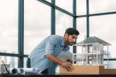 Ingénieur architecte caucasien professionnel inspecter le modèle de maison moderne avec vue sur le gratte-ciel tout en comparant le modèle de maison et le plan directeur avec le plan de projet et l'équipement architectural. Traçabilité.